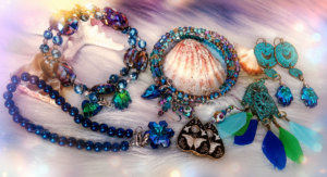 Handmade bohemian-style jewelry in hues of blue and green displayed with seashells on a soft white background.